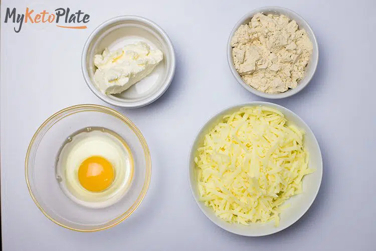 A top-down view of keto-friendly pizza ingredients: shredded mozzarella cheese, almond flour, cream cheese, and an egg, arranged in bowls.
