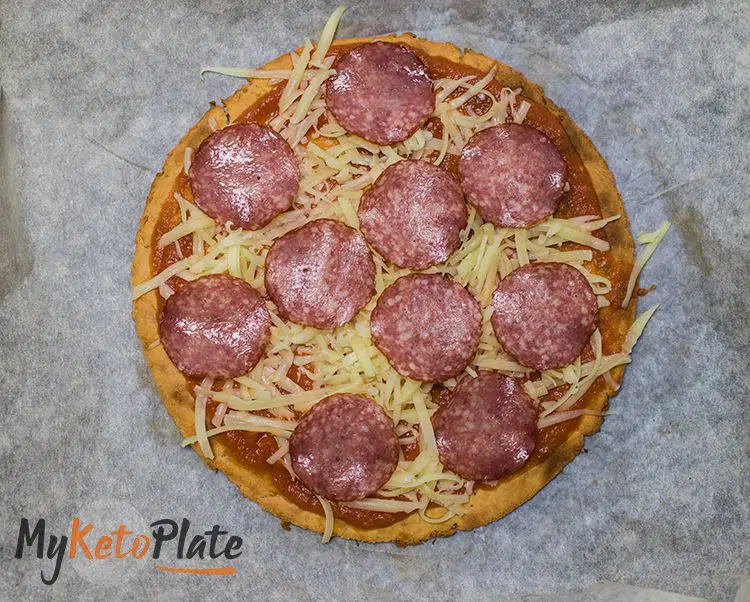 A top-down view of a prepared keto pizza before baking, with evenly spread toppings on a parchment-lined tray.