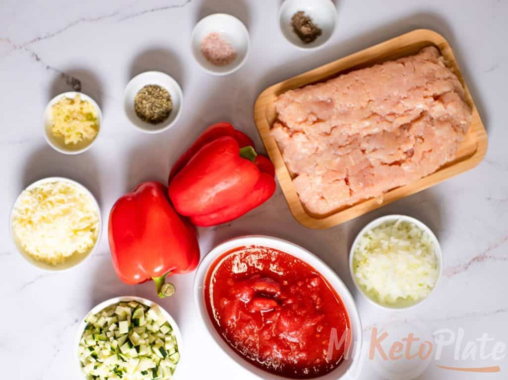 All ingredients for turkey stuffed peppers displayed on a white countertop, including bell peppers, ground turkey, zucchini, marinara sauce, and cheese.