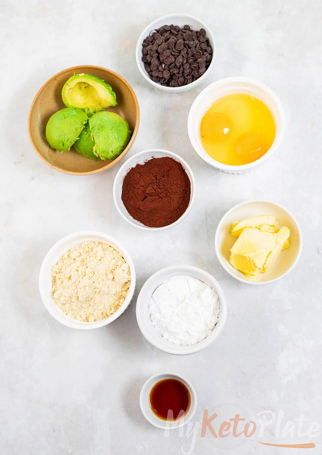 Top view of avocado brownie ingredients including avocado, cocoa powder, almond flour, eggs, and butter in small bowls.
