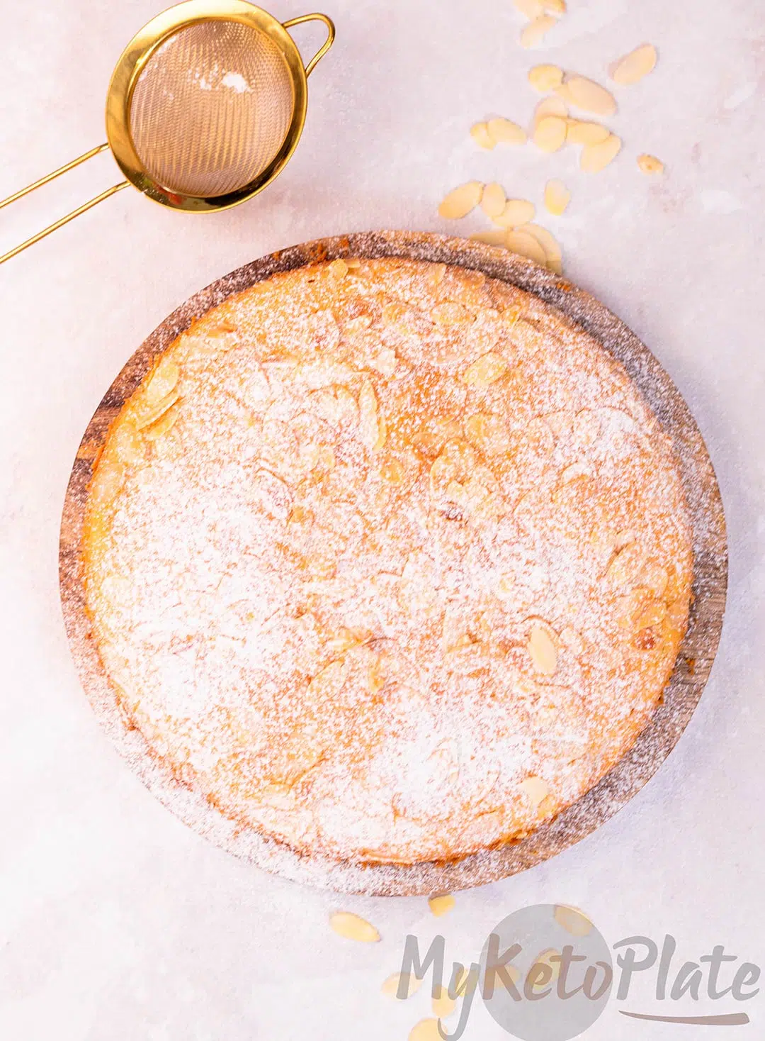 A golden-brown almond flour cake, freshly baked and dusted with powdered sugar, sitting on a wooden board.