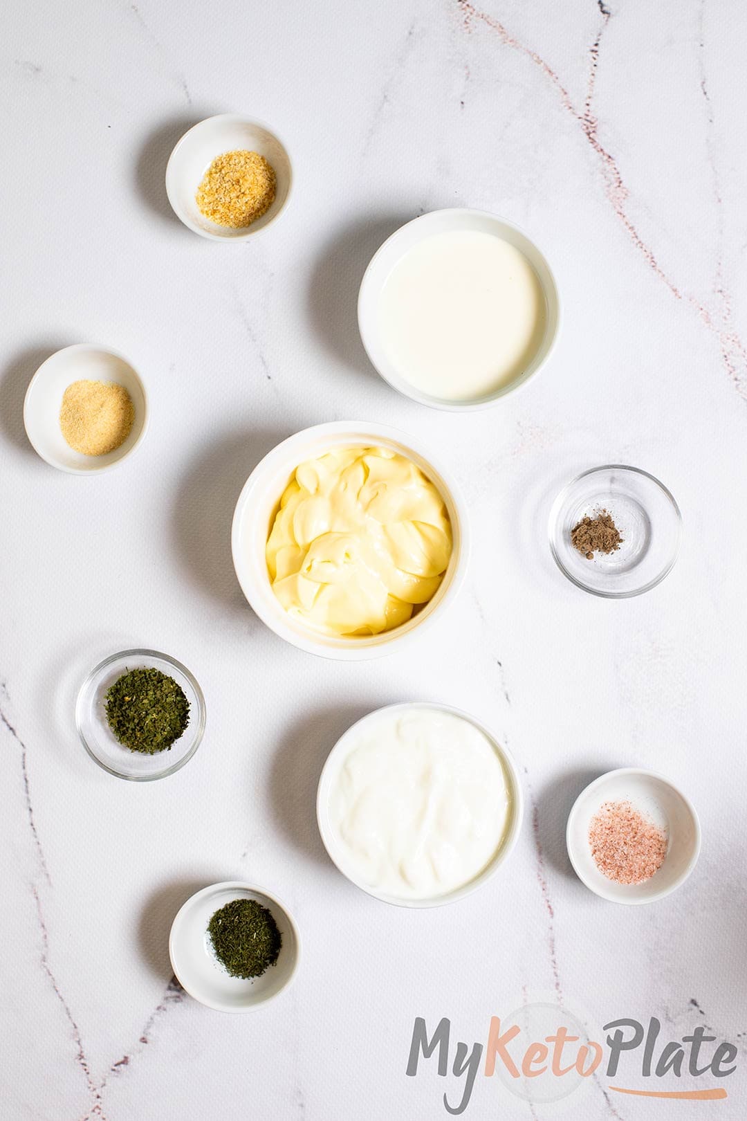 All the ingredients for homemade keto ranch dressing laid out on a marble countertop—mayonnaise, yogurt, herbs, and spices.