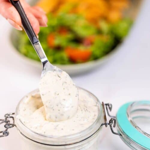 A spoon scooping thick, creamy keto ranch dressing from a glass jar.