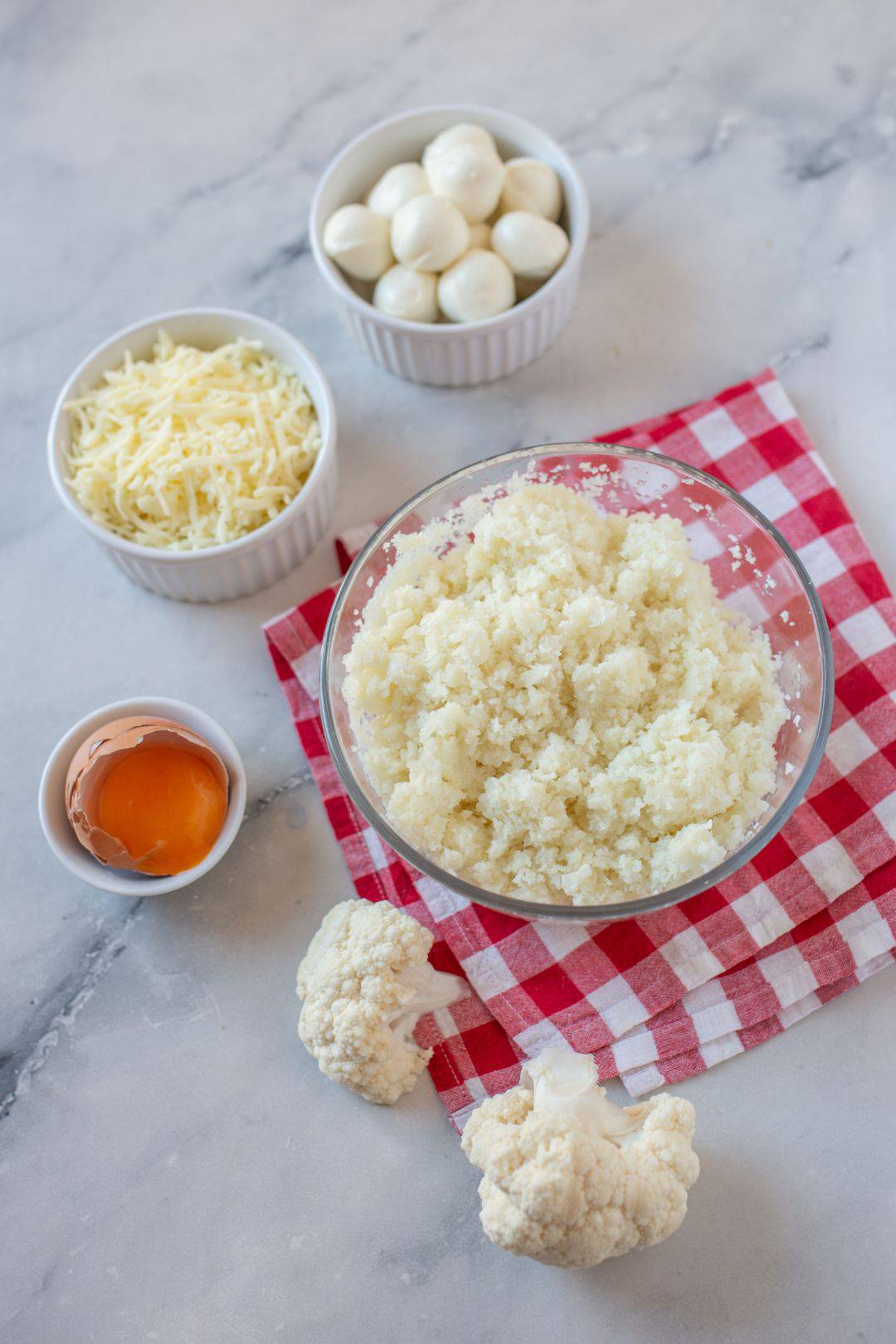 A top-down view of the ingredients for a cauliflower pizza crust, including riced cauliflower, shredded cheese, an egg, and fresh mozzarella.