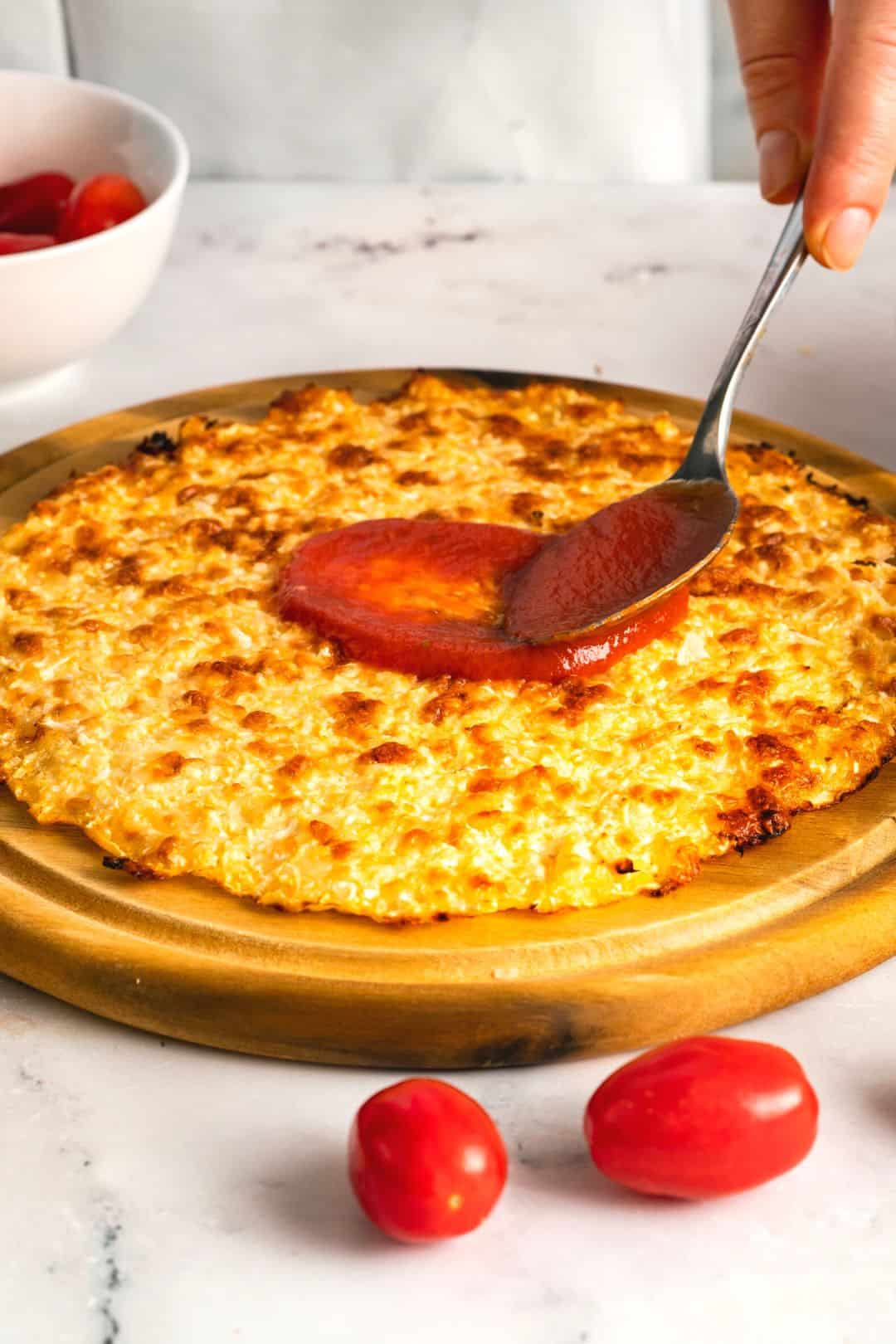 A perfectly baked cauliflower crust being prepared with a layer of fresh tomato sauce for a delicious low-carb pizza.