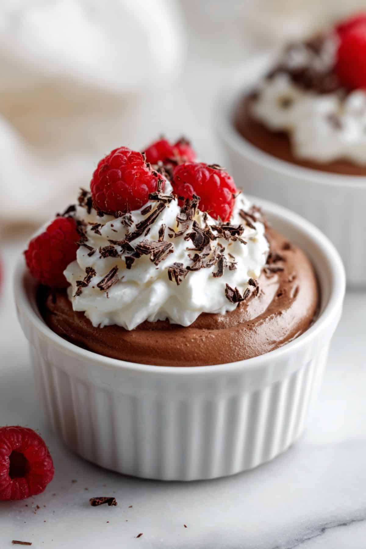 Elegant high-protein dessert in white ramekin, garnished with whipped cream peaks and three raspberries, shallow depth of field.