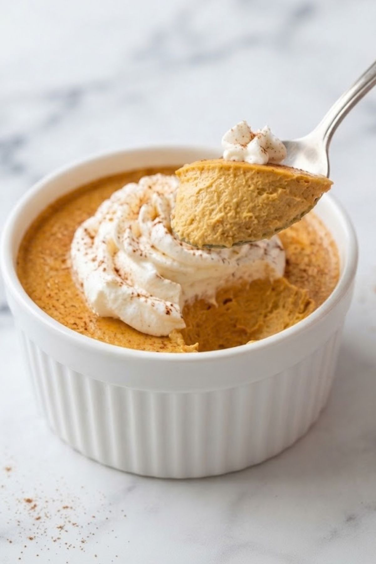 High-Protein Cottage Cheese Pumpkin Cheesecake 5 Close-up of High-Protein Cottage Cheese Pumpkin Cheesecake in a white ramekin, topped with whipped cream and cinnamon, with a spoon lifting a creamy bite.