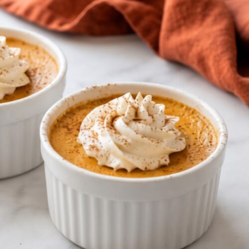 High-Protein Cottage Cheese Pumpkin Cheesecake 6 Two mini High-Protein Cottage Cheese Pumpkin Cheesecakes in white ramekins, topped with whipped cream and cinnamon on a marble surface.